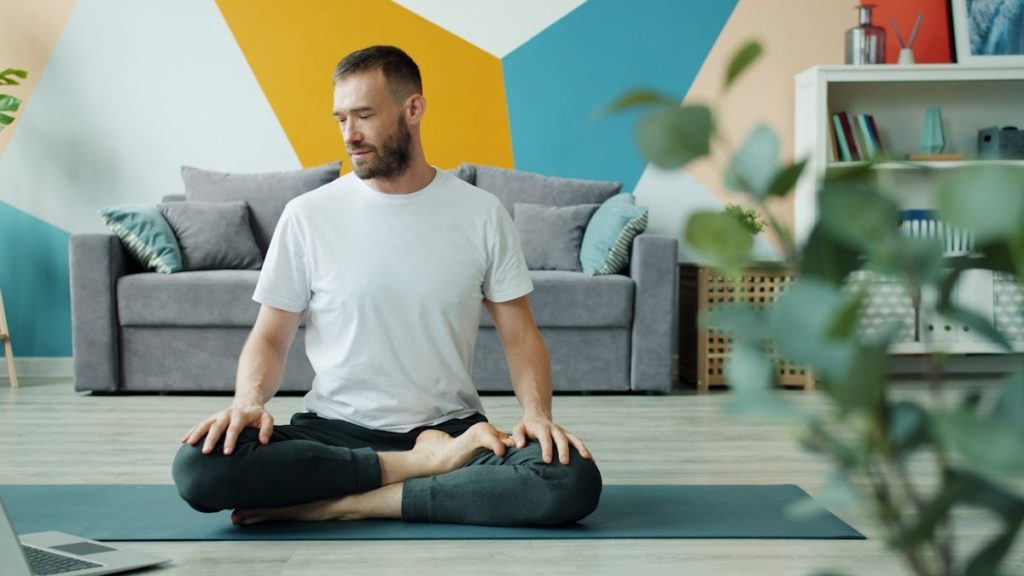 Yoga student serious man is learning asanas online using laptop at home exercising on mat in cozy apartment using portable computer. People and house concept.
